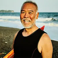 Happy senior man with graying hair smiling on a beach with ocean waves and a coastline in the background.