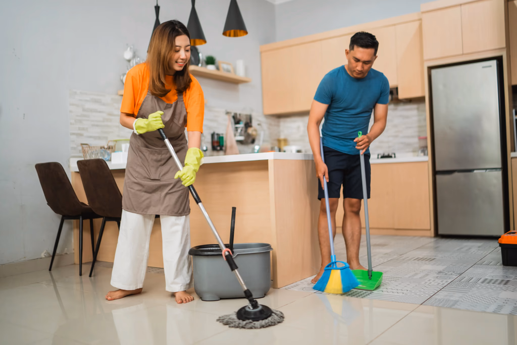 A young couple working together to clean their home kitchen using a mop, broom, and bucket.