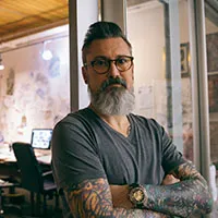 Portrait of a middle-aged man with a gray beard and tattoos standing in a modern office or studio setting.