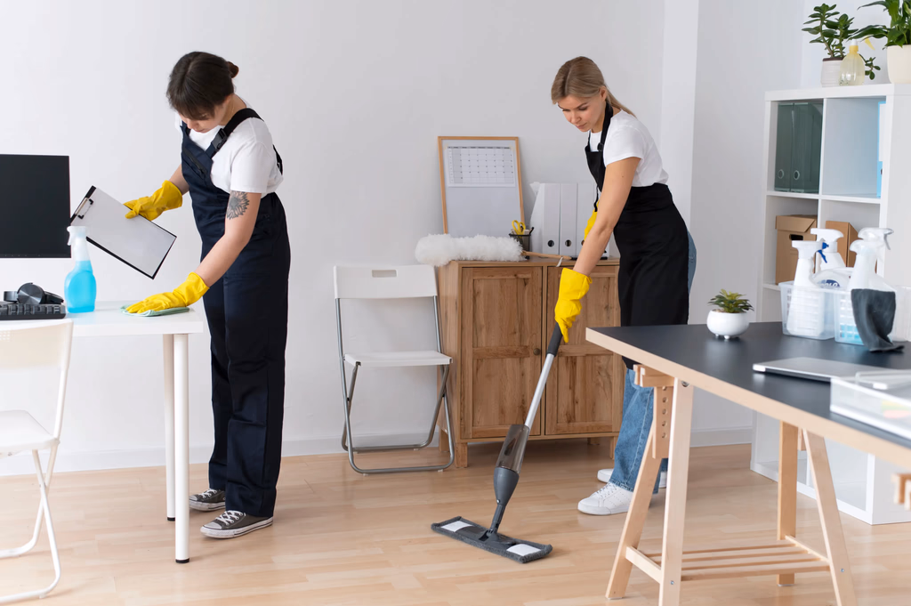 Two professional cleaners in black aprons and yellow gloves deep cleaning an office space with a mop and microfiber cloth.